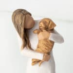 Standing female figure in cream dress, holding golden color puppy in her arms